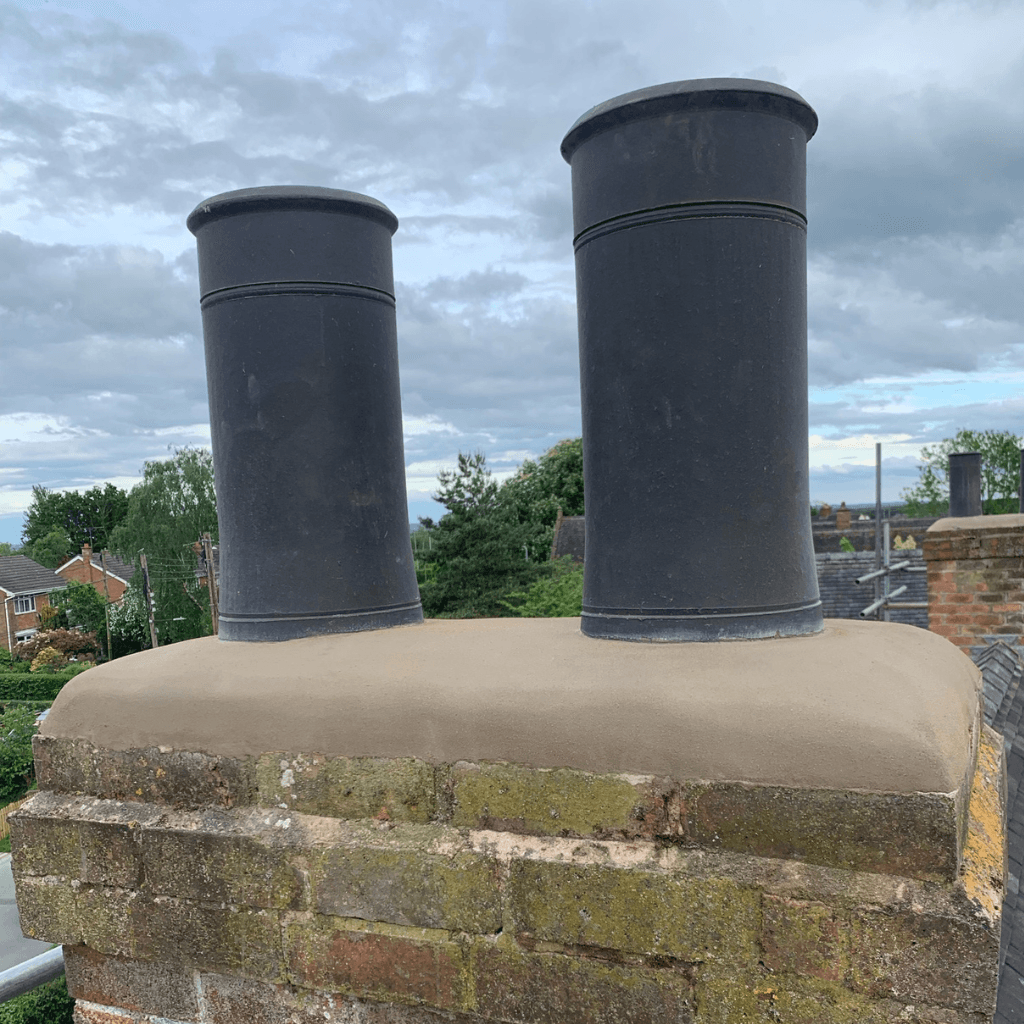 Close-up of two dark grey cylindrical chimney pots set into a fresh, smooth cement mortar crown on an old red brick chimney stack, overlooking a village with green trees and a cloudy sky.