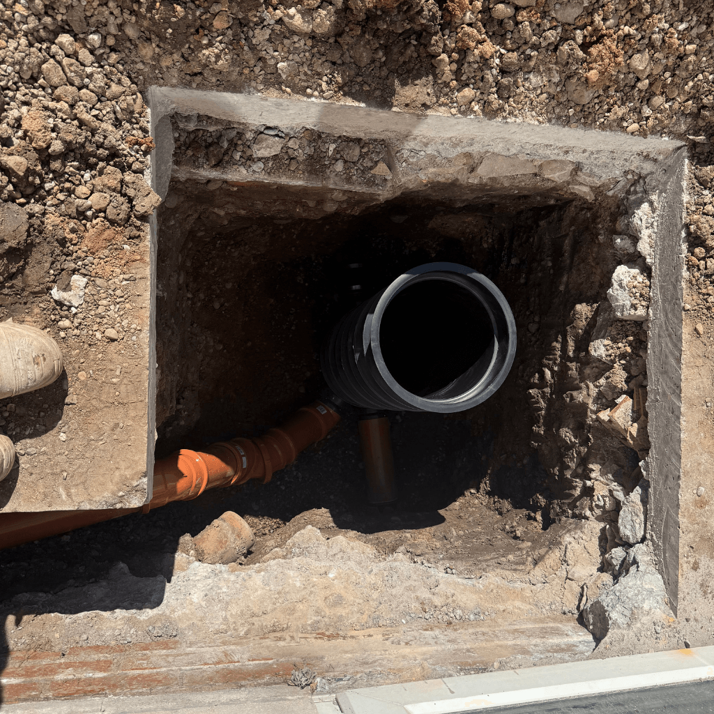 Top-down view into a square excavation showing a black plastic inspection chamber connected to several orange underground drainage pipes in a dirt and gravel trench.