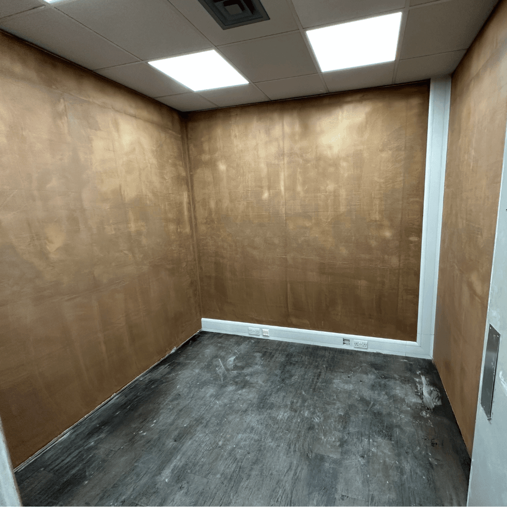 A room with textured golden walls, unpolished floor, and recessed electrical outlets. The room is partially furnished with ceiling lights overhead.