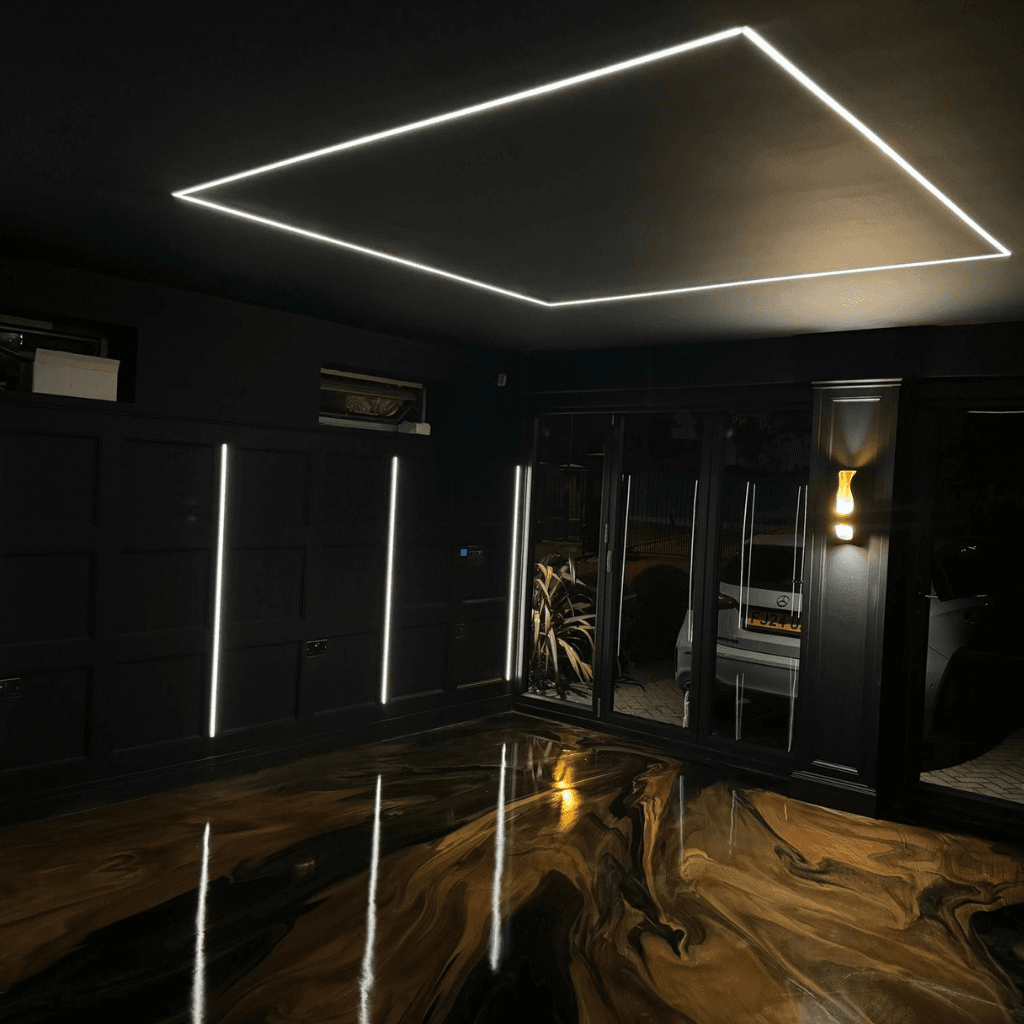 Modern black garage or studio with a high-gloss black and gold marble-effect epoxy floor. The room features a large square LED ceiling light, vertical LED strip lights on the walls, and glass doors reflecting the glowing interior