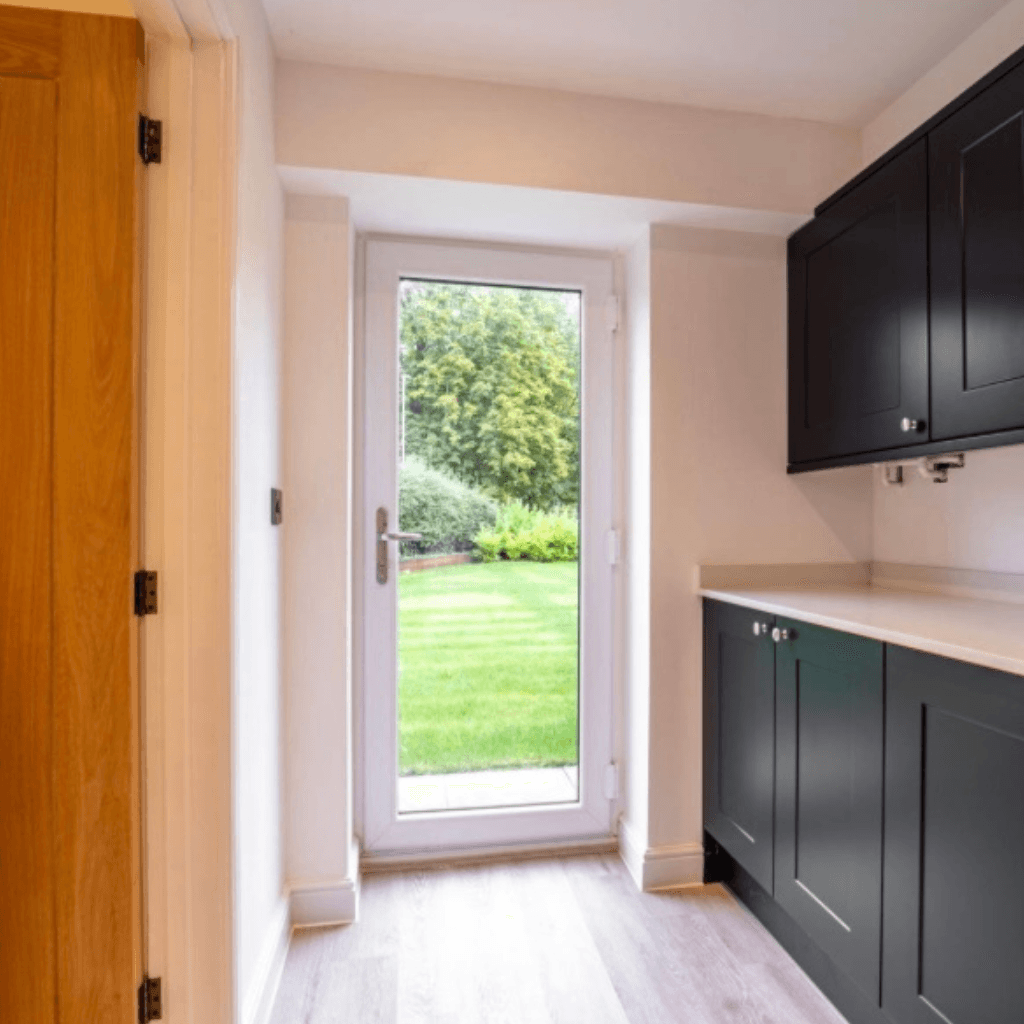A modern utility room with dark charcoal cabinets, a light stone countertop, and a white uPVC glass door leading to a lush green lawn. The room has light wood-effect flooring and a natural wood door on the left.