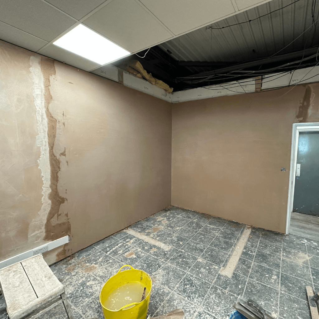 "A room with unfinished brown plaster walls, a bucket of water on the floor, and exposed ceiling beams with insulation. Part of the floor tiles are incomplete, and a door is visible on the right side.