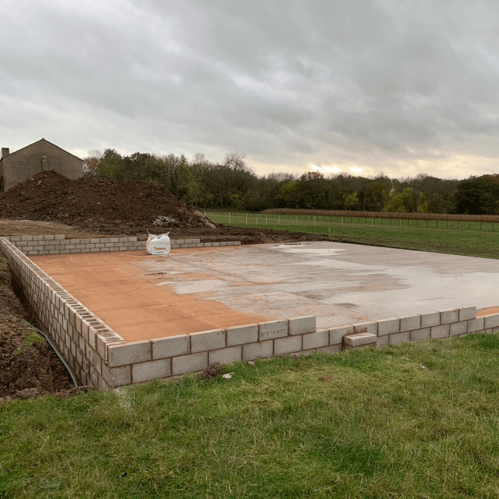 A large rectangular building foundation in a grassy field with a low perimeter wall made of grey cinder blocks. The interior is partially filled with a layer of orange sand and wet grey concrete under an overcast sky.