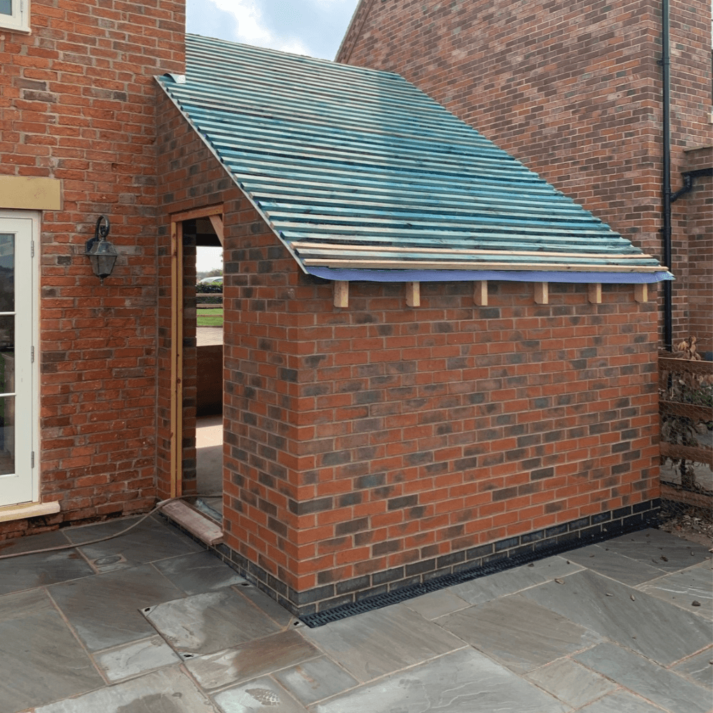 A small red-brick house extension with a sloped roof frame featuring blue horizontal battens and a visible roofing membrane. The structure is built onto a stone-paved patio next to an existing two-story brick home.