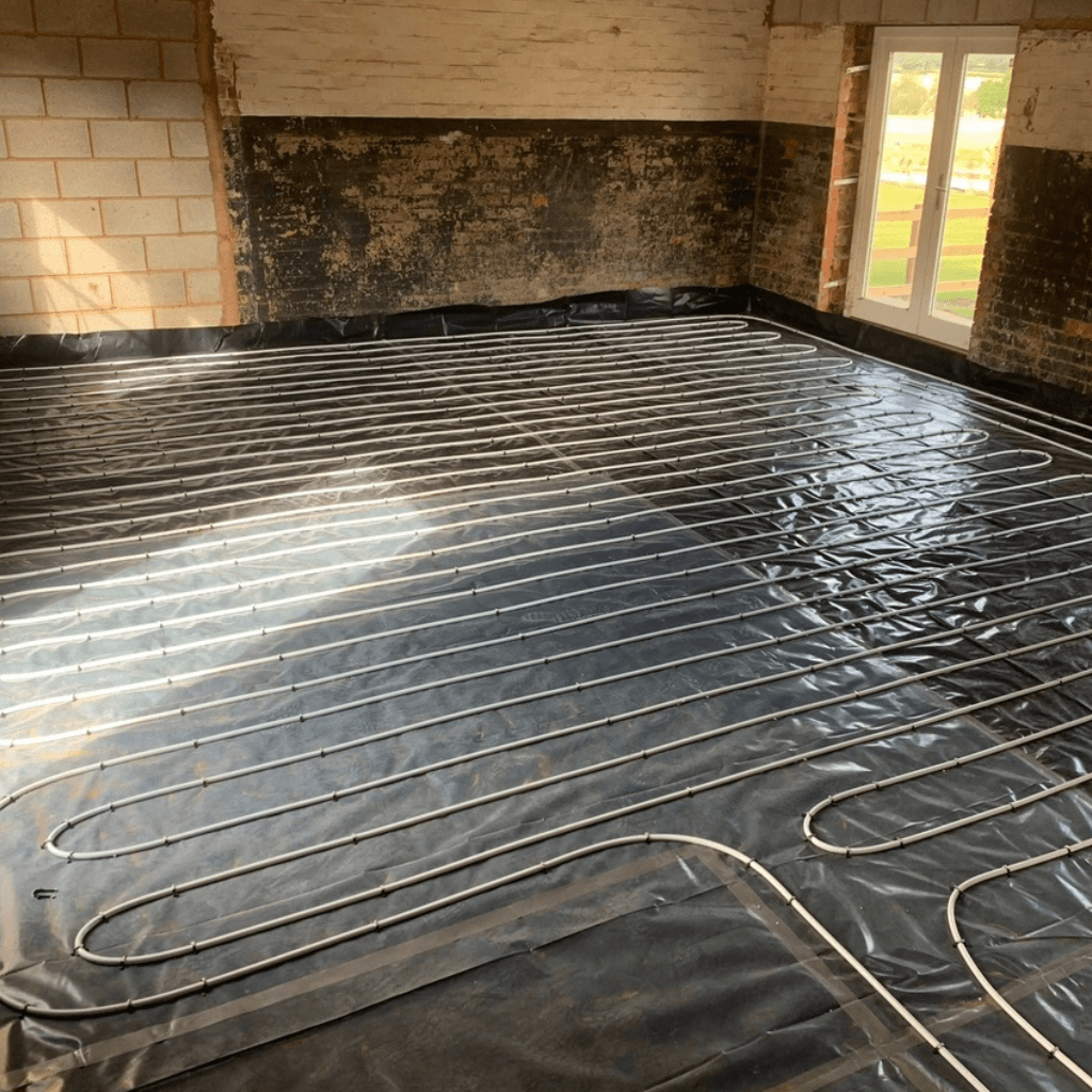 Interior room under renovation featuring a wet underfloor heating system with white piping laid in a serpentine pattern over a black plastic membrane. The room has exposed brick and block walls and a white patio door.