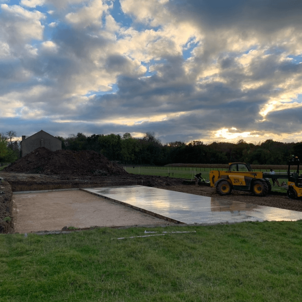A construction site in a grassy field at sunset, showing a large concrete slab foundation partially poured next to a section of orange sand. A yellow JCB telehandler is parked nearby against a backdrop of trees and a cloudy sky.