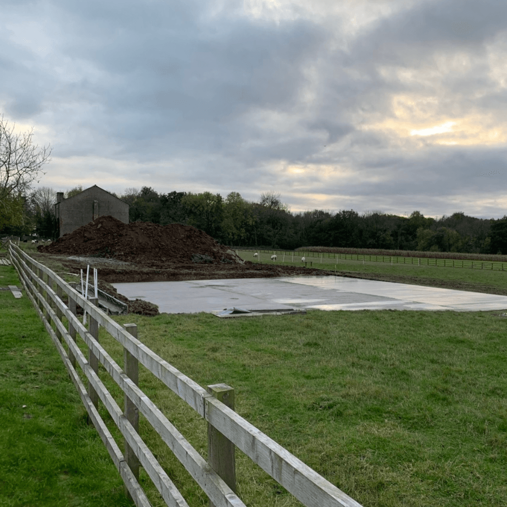 A large rectangular concrete slab foundation poured in a grassy rural field. A tall mound of brown earth sits behind the foundation, with a wooden post-and-rail fence in the foreground and a cloudy sky above.