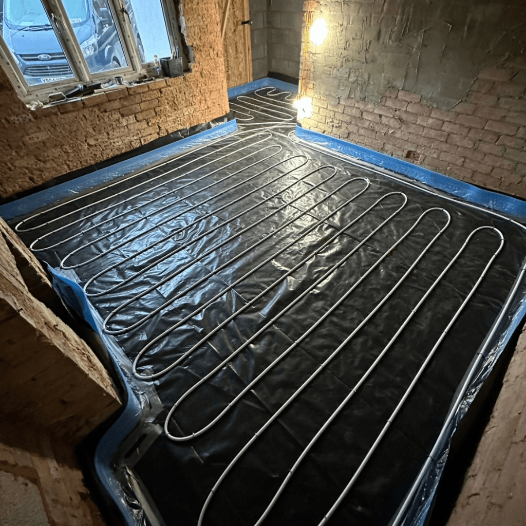 A floor heating system installation in progress, showing coiled pipes on a black membrane inside an unfinished room with exposed brick walls and large windows.