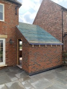 A small brick house extension in progress, showing a sloped roof frame with blue roofing membrane and horizontal wooden battens, attached to a two-story red brick home with a stone patio.